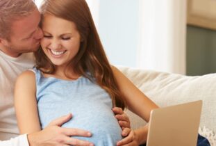 זוג בהריון יושבים ביחד על הספה עם לפטופ ומחייכים